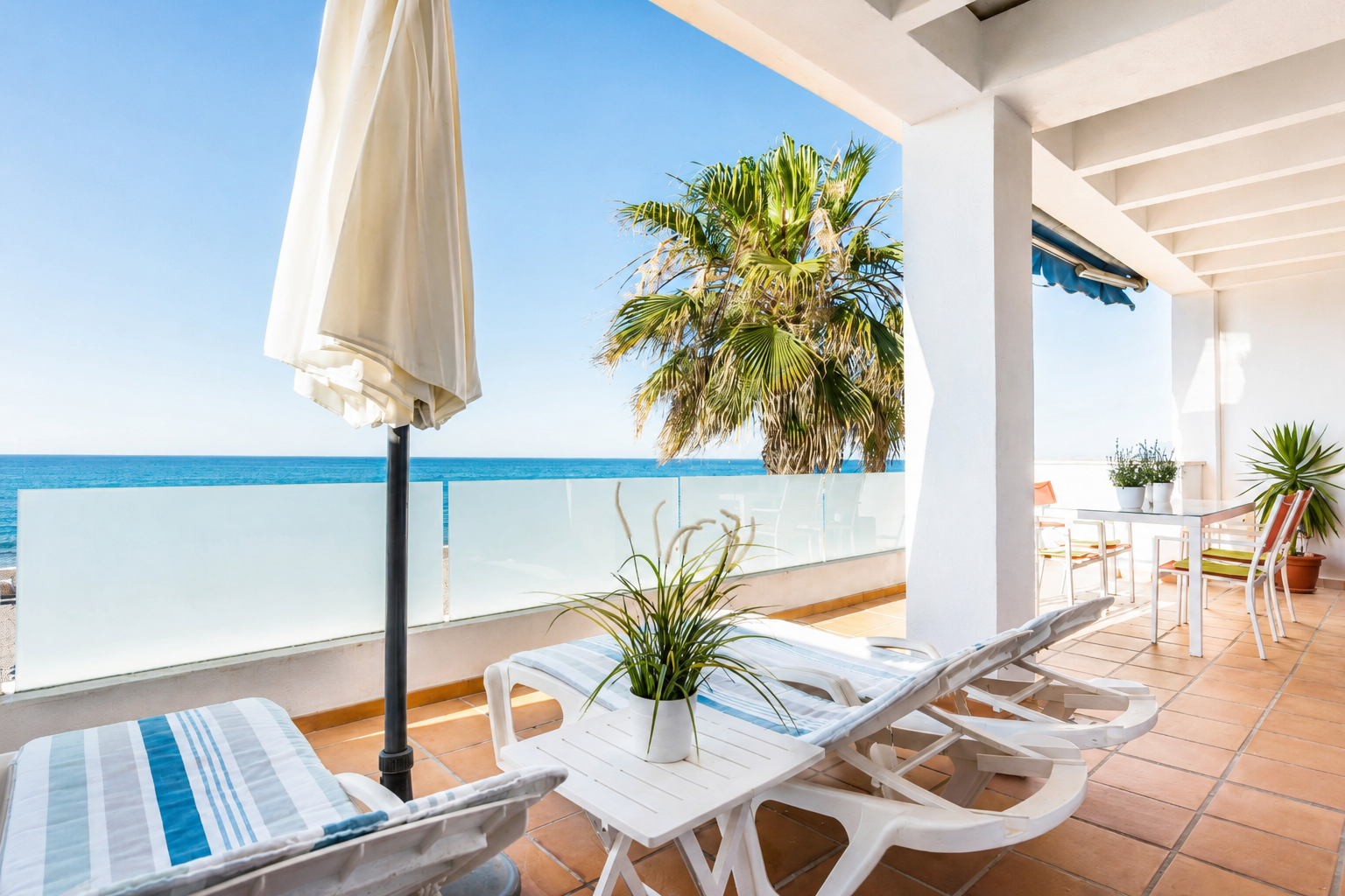 Terraza junto al mar con tumbonas blancas, mesa, sombrilla y palmera frente a cielo azul.