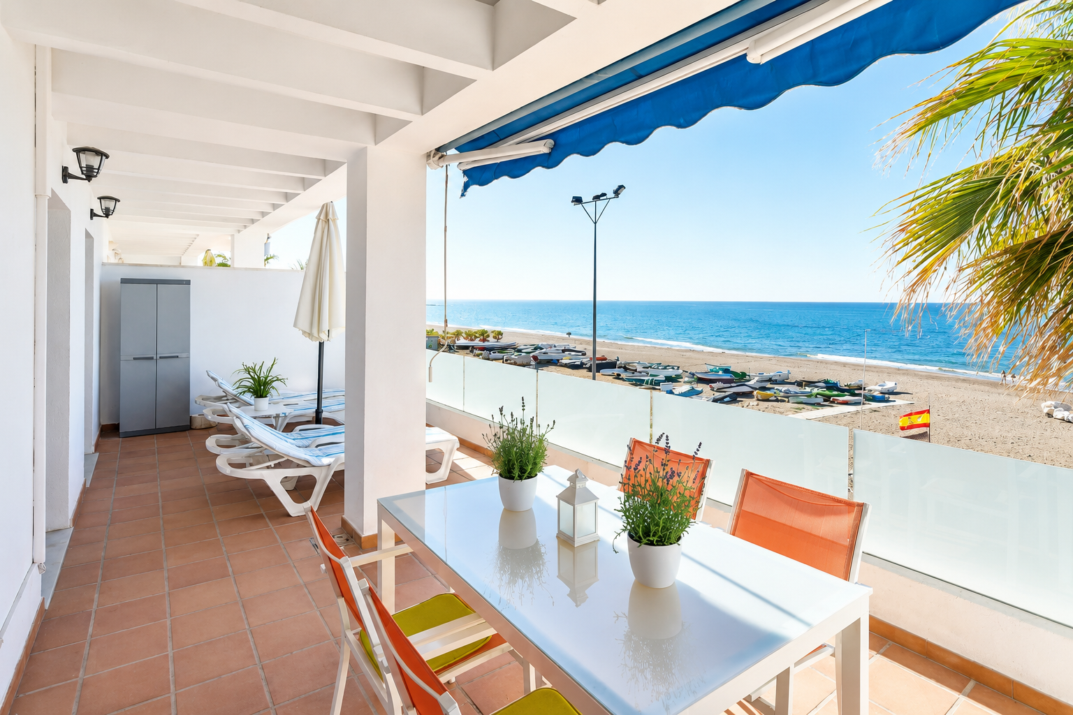 Terraza junto al mar con tumbonas blancas, mesa, sombrilla y palmera frente a cielo azul.