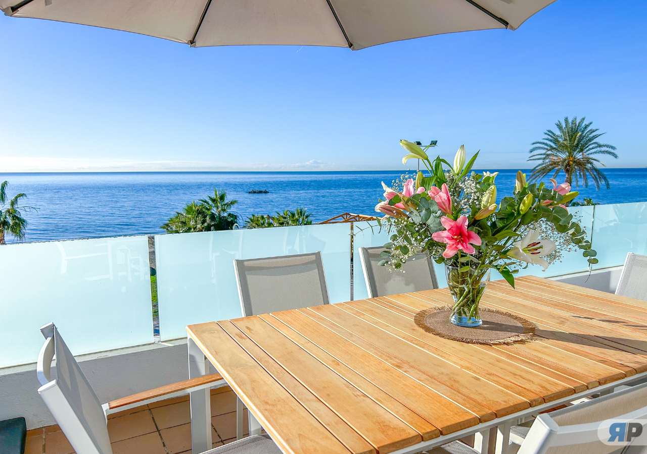 Mesa de madera con jarrón de flores bajo sombrilla, sillas blancas y vista al mar azul.
