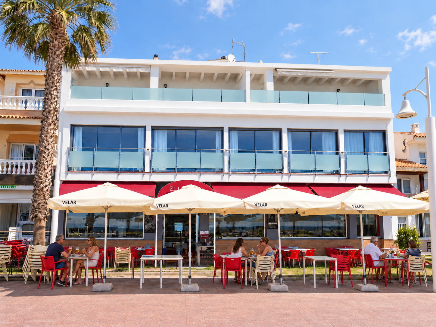 Terraza de hotel con mesas y sillas rojas, sombrillas blancas y fachada moderna bajo cielo azul.