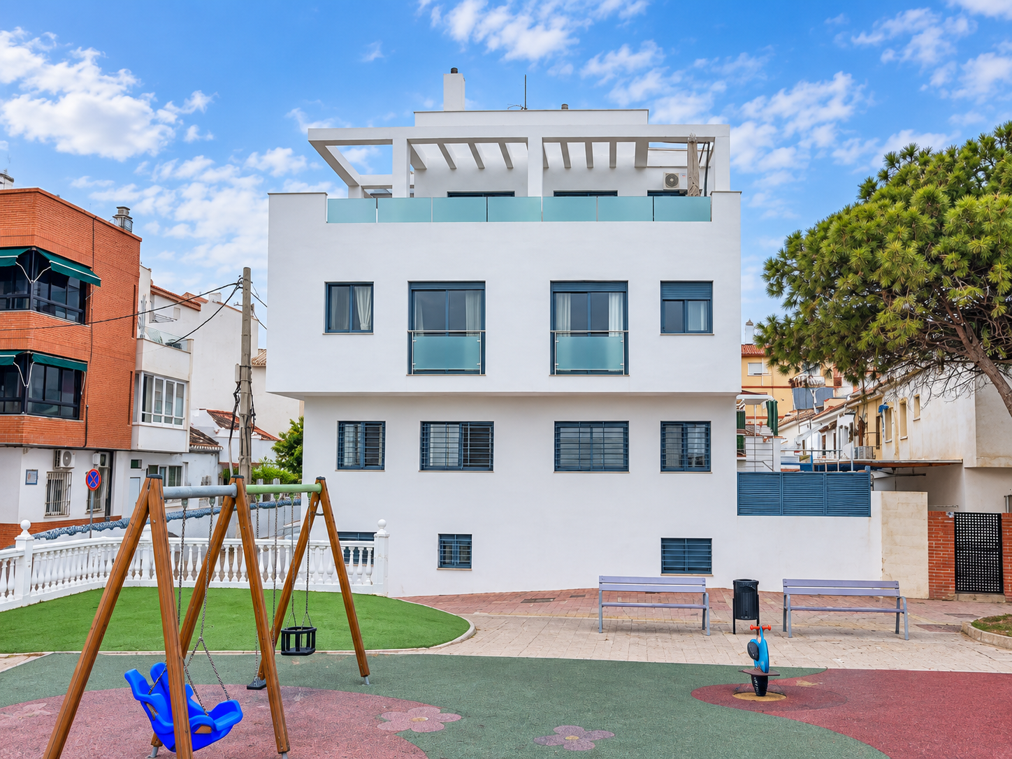 Fachada blanca moderna con ventanas azules, patio con columpio infantil y cielo azul con nubes.